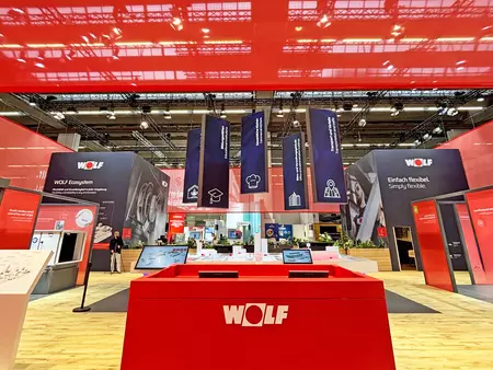 Messestand von WOLF mit zentralem roten Infocounter, großem roten Deckenelement, hängenden Bannern und Produktbereichen im Hintergrund.